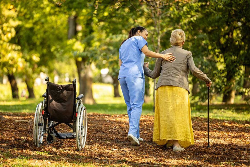 An adult daughter holds hands with her elderly mother in a sunlit living room.